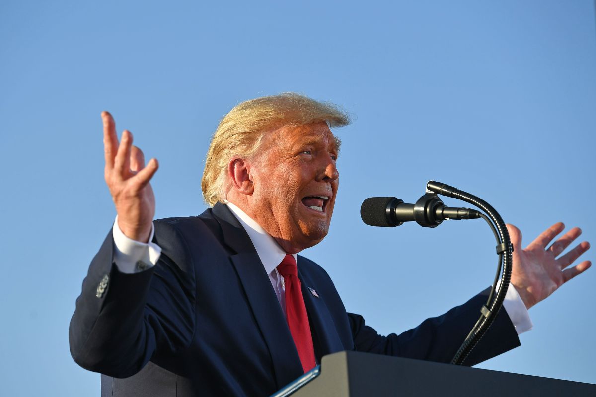 Donald Trump speaking into a microphone against a backdrop of the sky. He is gesticulating with his hands.
