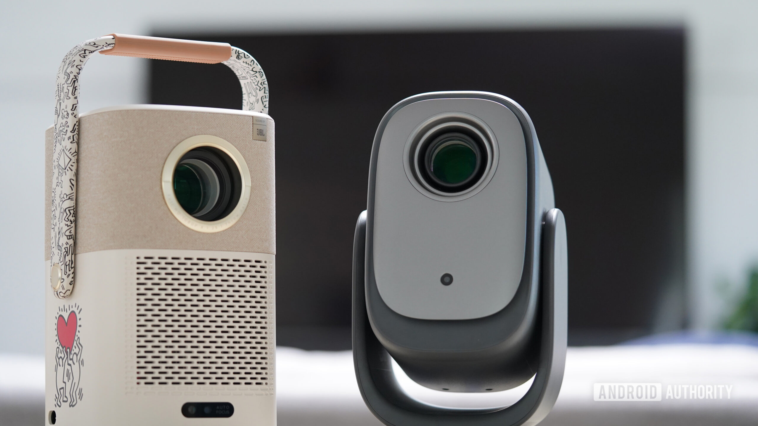Two projectors stand in front of a large TV.